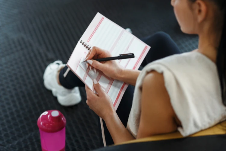 Zdjęcie siedzącej kobiety w sportowym ubraniu, notującej w zeszycie z planem tygodnia.