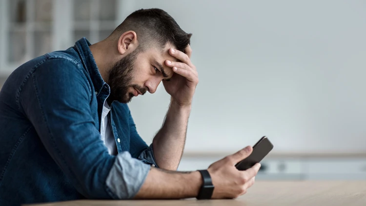Mężczyzna siedzący przy stole podpierający głowę ręką i patrzący w telefon