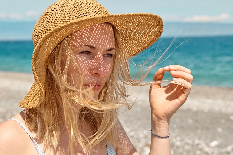 Portret blondynki w słomkowym kapeluszu plażowym, zasłaniającej dłonią twarz przed ostrym, letnim słońcem