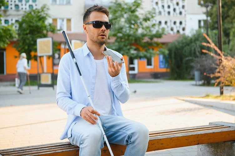 Mężczyzna z białą laską korzysta z telefonu na ławce w przestrzeni miejskiej